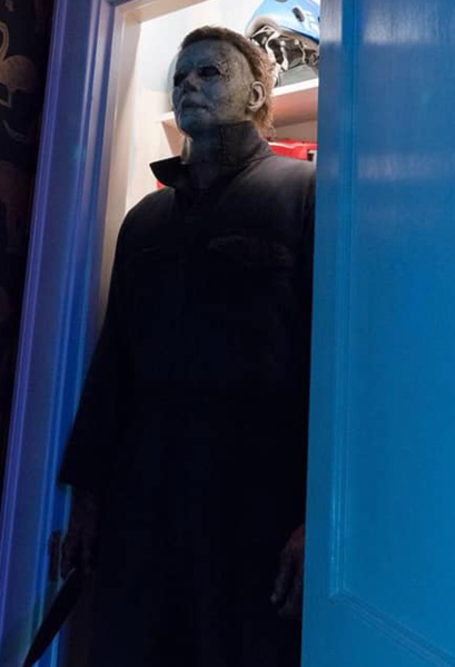 This is a Halloween 2018 Michael Myers mask that is a weathered grey face, neck and ear and has brown hair and black eyes and he is wearing grey coveralls, holding a knife and standing in a doorway.
