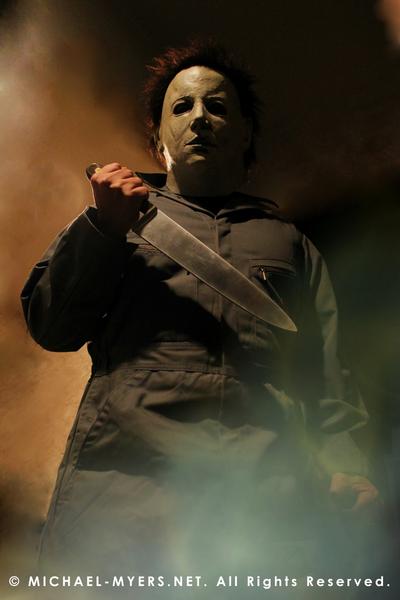 This is a Halloween 6 Curse of Michael Myers mask that is white with brown hair and black eyes and he is wearing coveralls and standing in fire and holding a knife..