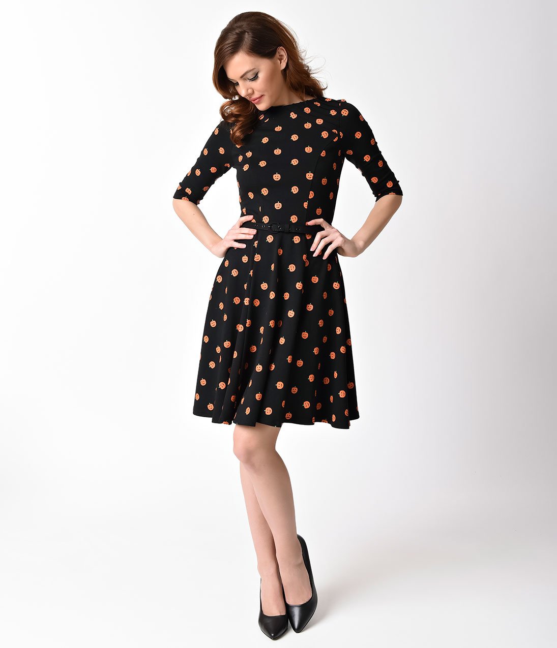 This is a black pinup flare dress with orange pumpkins, belt and 3/4 sleeves and the model is wearing black shoes and has brown hair.