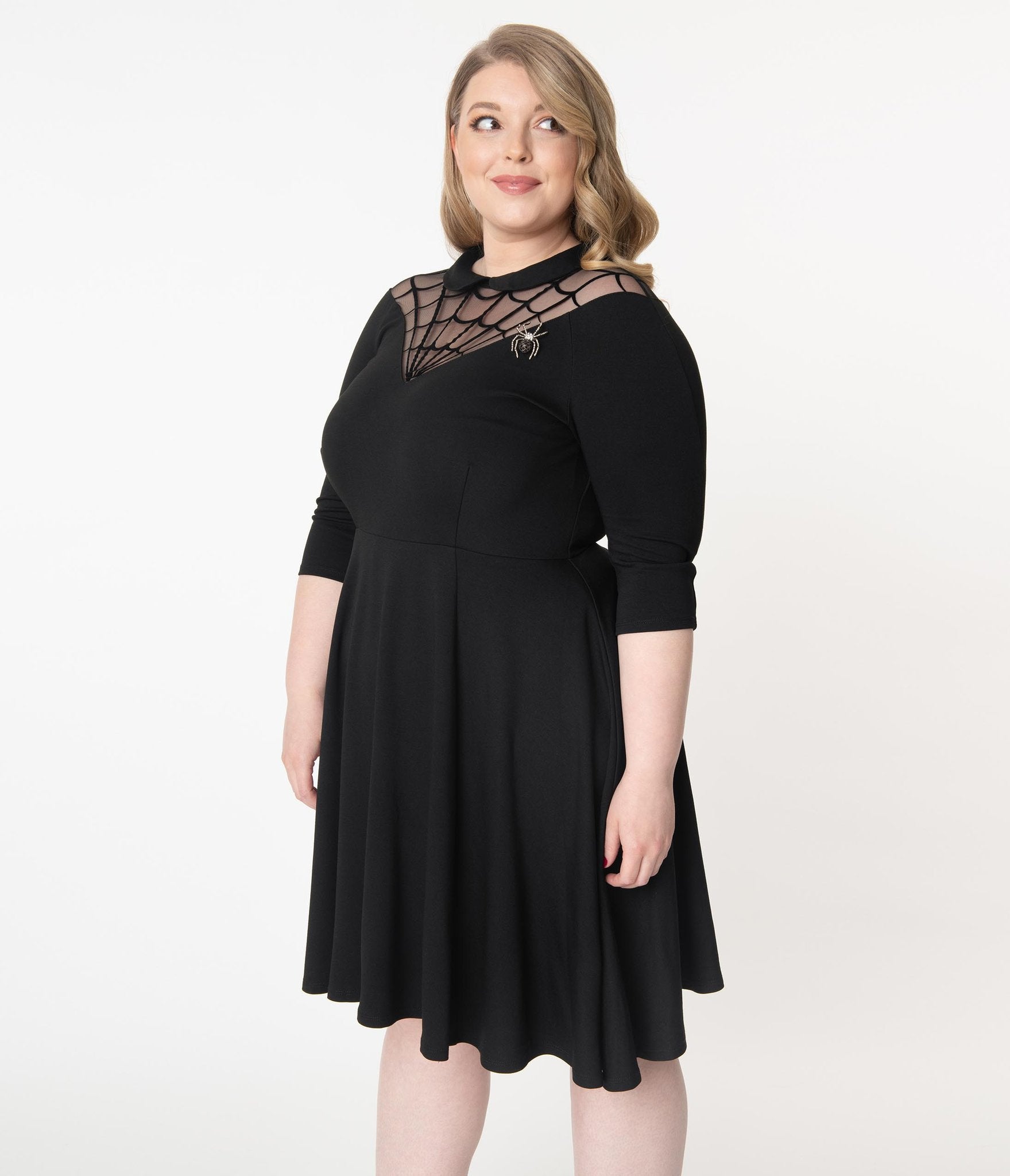 This is a black Unique Vintage flare dress that has a spiderweb neck, Peter Pan collar, 3/4 sleeves and the plus model is smiling and has a spider brooch.