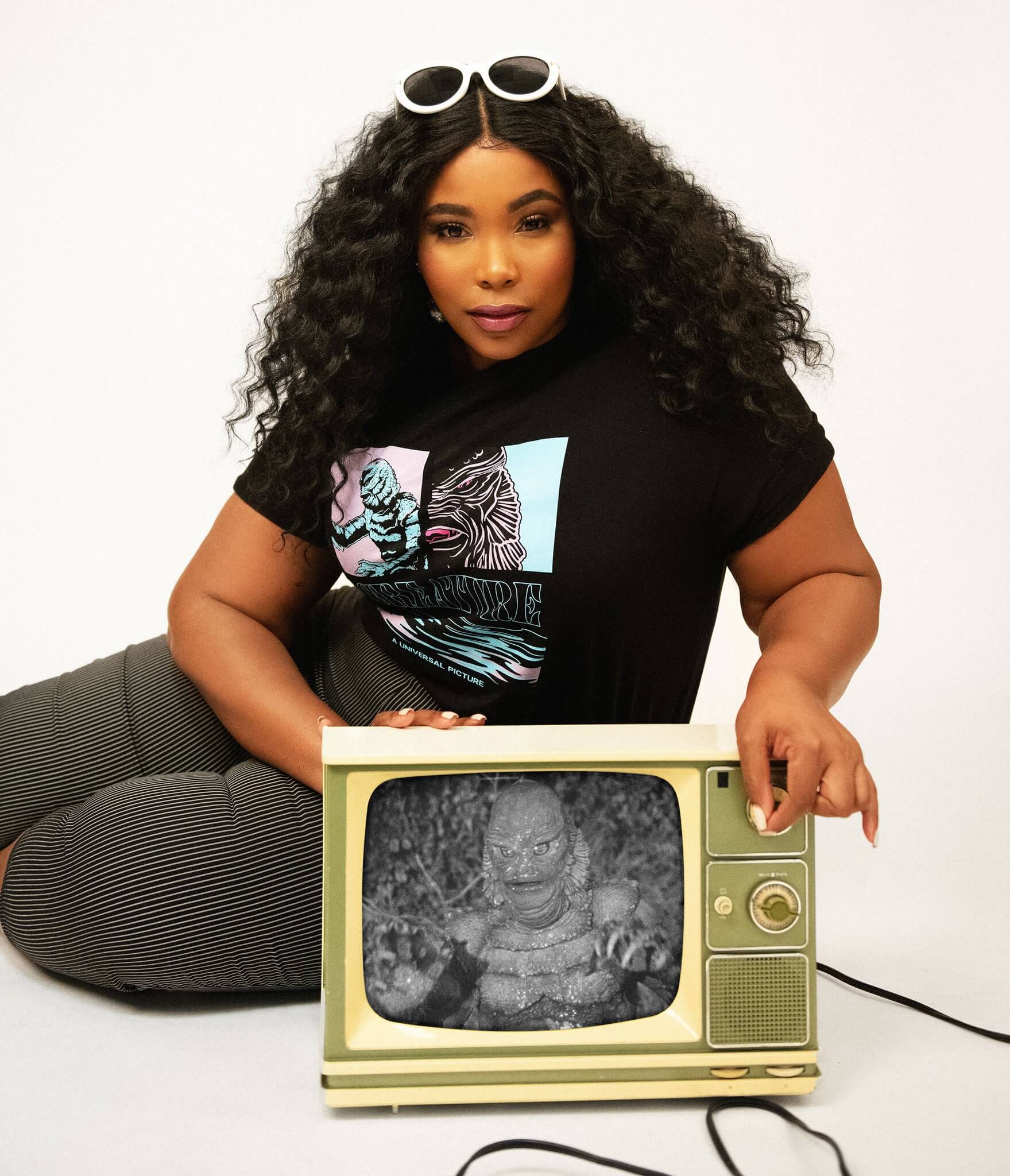 This is a Universal Monsters Creature From the Black Lagoon Unisex Tshirt by Unique Vintage and it is black with pink and blue graphic and creature has gills and scales and the model is a girl with dark hair and she has on black pants and is touching a green TV.