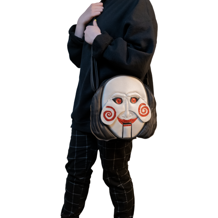 This is a Saw Jigsaw Billy purse and he has a white face, red eyes, red lips, red spiral cheeks and black hair and the person wearing it has on black pants and a black sweatshirt.