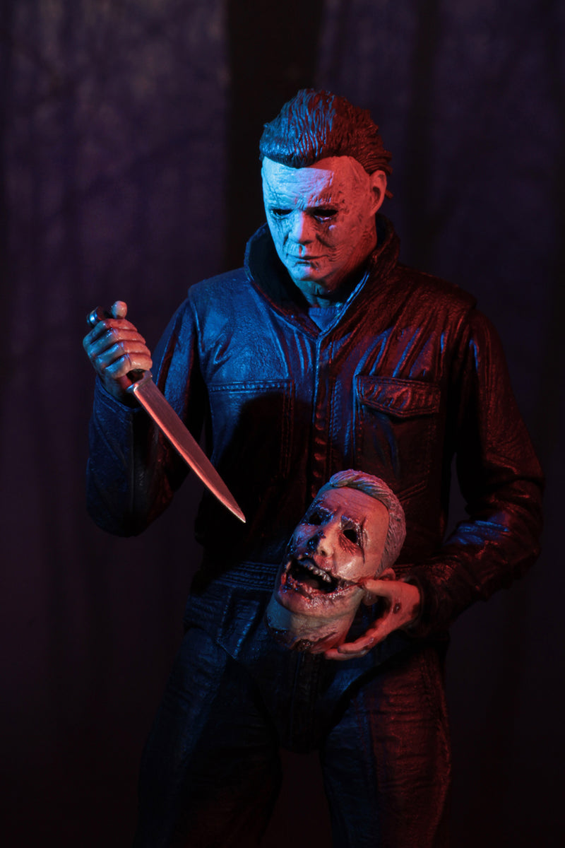 This is a Halloween 2018 Michael Myers NECA 7" ultimate action figure and he has a white mask, grey coveralls and black boots and he is holding a knife and a severed head.