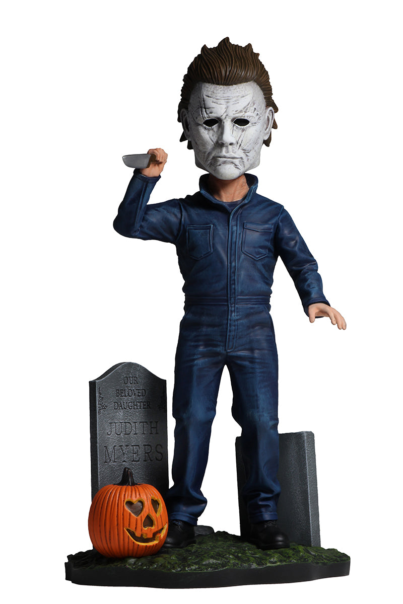 This is a 2018 Michael Myers NECA head knocker, where he is holding up a knife in his hand, has tombstones and a pumpkin around him and is wearing a white mask with brown hair and grey coveralls.