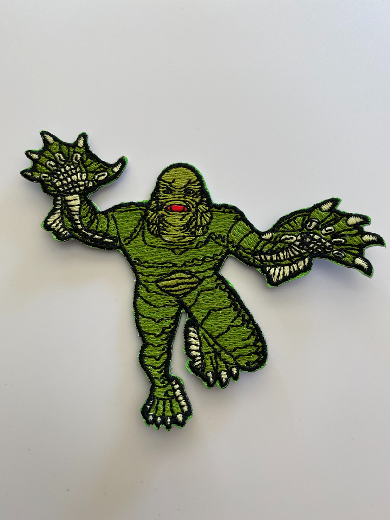 This is a Universal Monsters Creature From the Black Lagoon patch and he has a green face, black eyes and gills on the side of his head, webbed feet and hands and scales on his body.