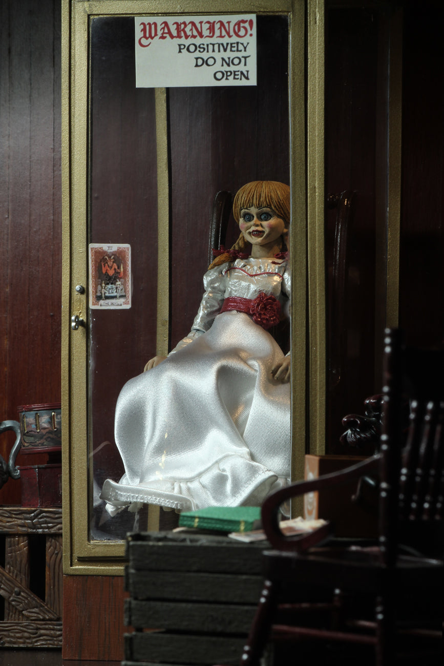 Annabelle NECA action figure from the Conjuring is sitting in a glass display case in a museum, on a rocking chair and has a white dress white a red sash and braids in her hair..