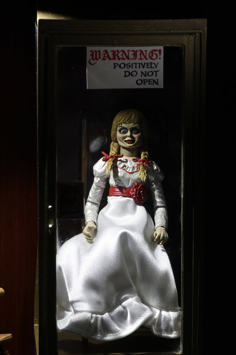 Annabelle NECA action figure from the Conjuring is sitting on a rocking chair in the Warren occult museum and has a white dress white a red sash and braids in her hair.