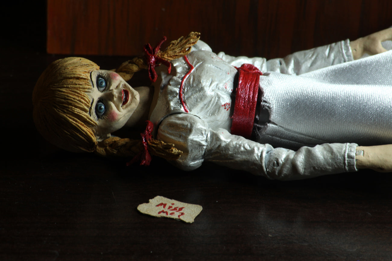 Annabelle NECA action figure from the Conjuring is laying down in a white dress with a red sash, next to a note she wrote in red letters that says miss me..