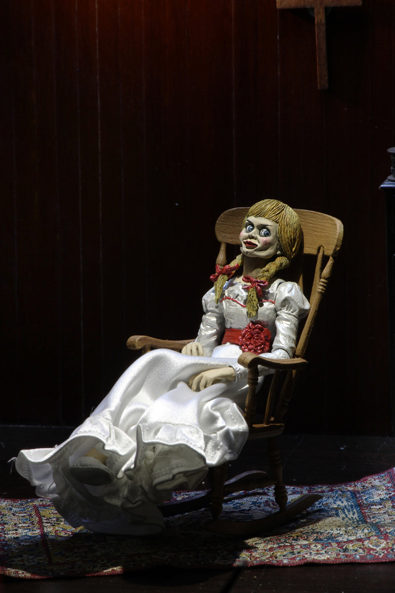 Annabelle NECA action figure from the Conjuring is sitting on a rocking chair and has a white dress white a red sash and braids in her hair..