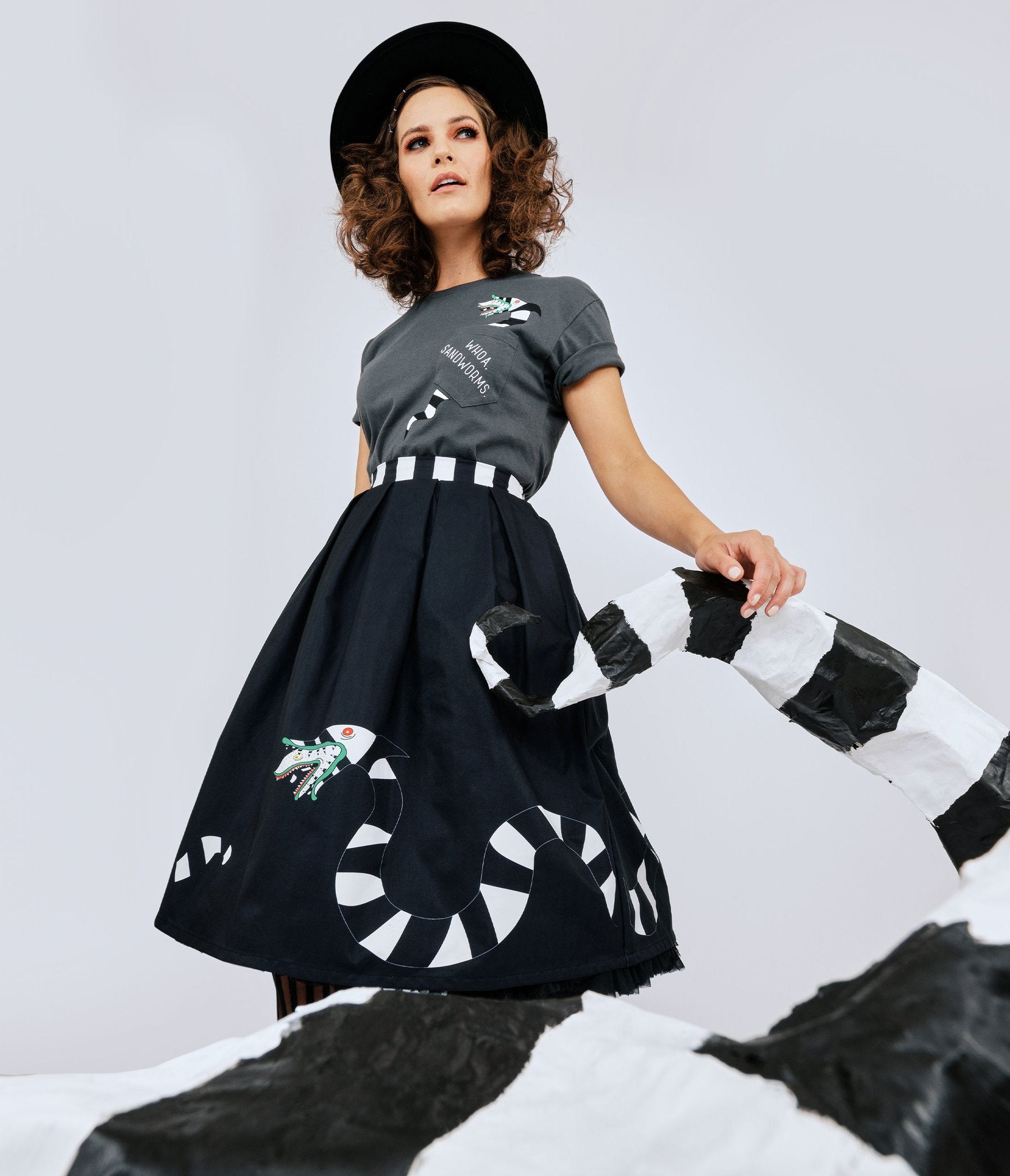 This is a Beetlejuice vintage pinup Sandworm swing skirt from Unique Vintage, that is black with a striped worm and the model is wearing a grey shirt and black hat.