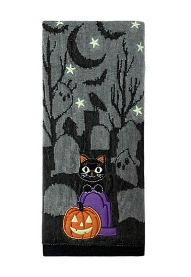 This is a grey hand towel and it has a black cat on a purple headstone in a cemetery, with an orange pumpkin, ghosts, bats crows, moons and glowing stars.