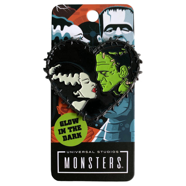 This is a Universal Monsters Bride of Frankenstein Heart enamel pin and has a woman with a white face, black hair that has a white streak and red lips and a greek man with black hair and stitches in his face.