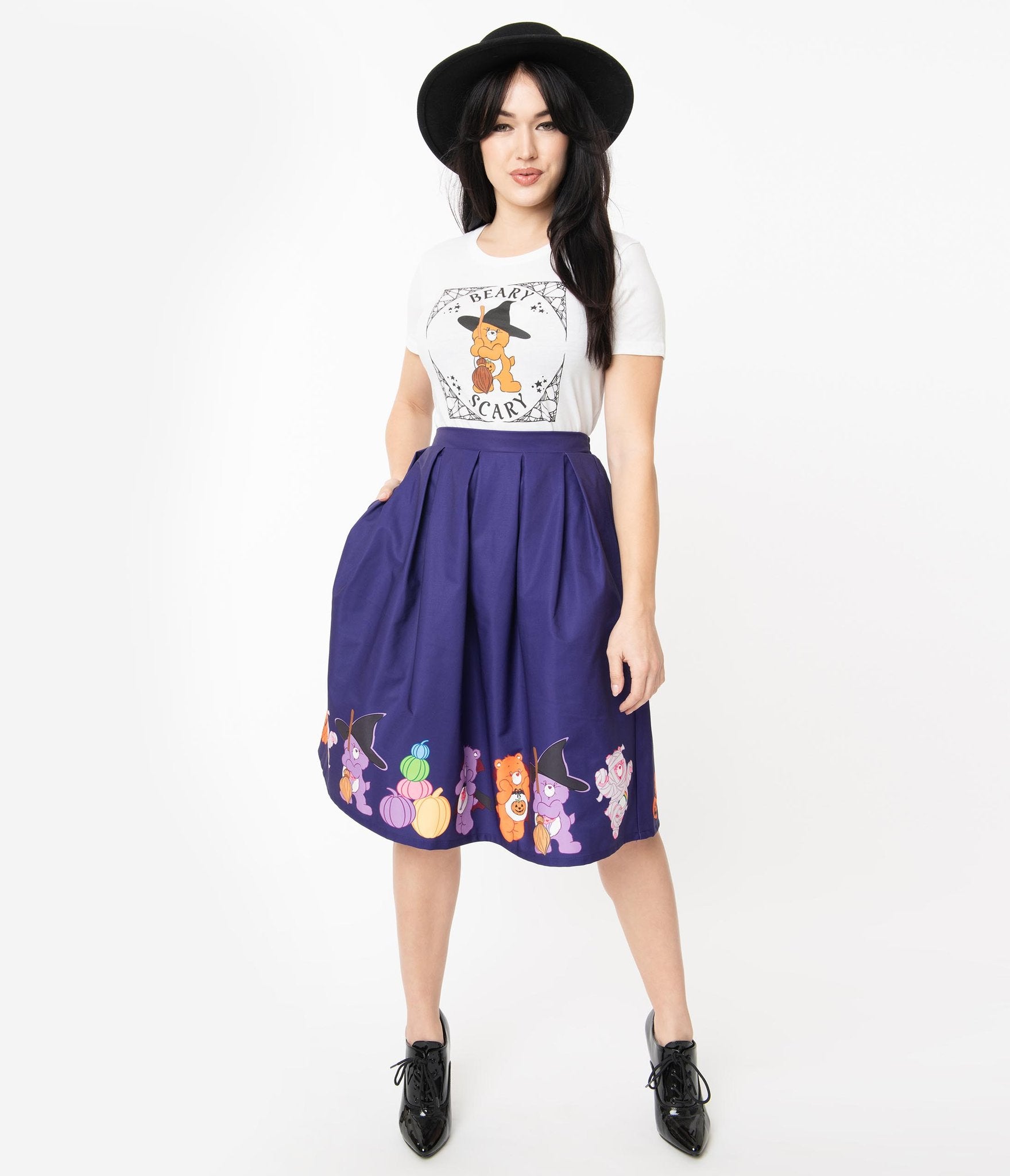 This is a Care Bears halloween purple Jayne swing skirt by Unique Vintage and the model is wearing a white shirt and has on black shoes and a black hat and her hand is in the pocket. 