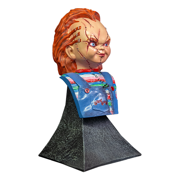 This is a Bride of Chucky mini bust and he has orange hair, a striped shirt, blue overalls and stitches on his face, has hair stapled to his head and is on a grey stand.