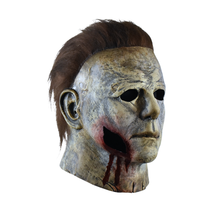 This is a weathered Michael Myers mask bloody edition from Halloween 2018 and it is a grey and brown face and ear with brown hair and black eyes and has a piece of face missing with blood coming out..