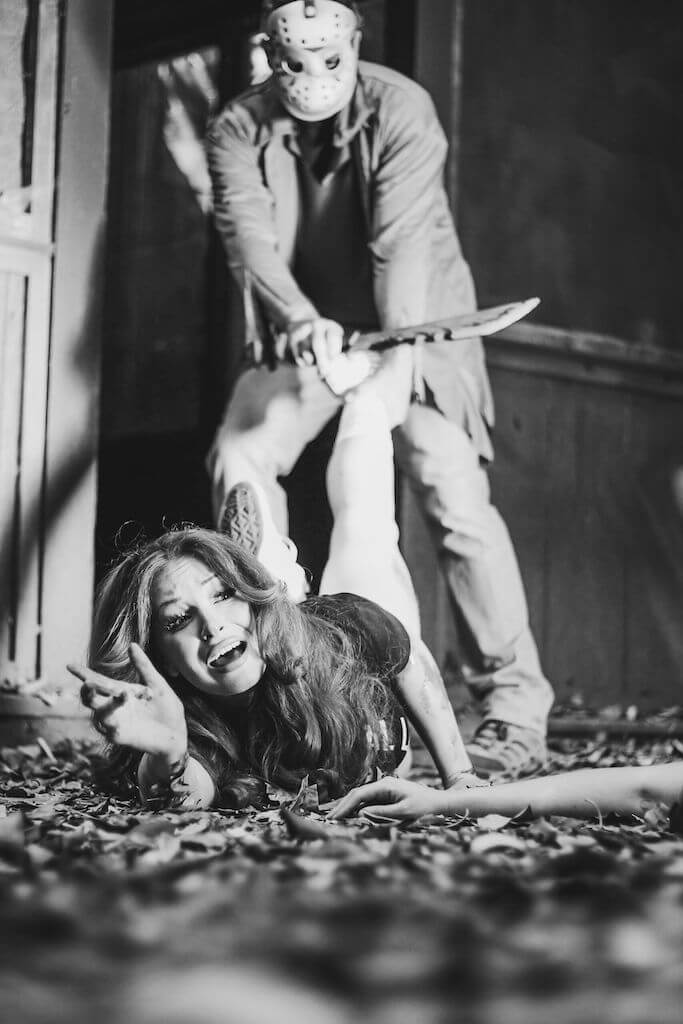 There is a camp counselor with red hair, a black camp crystal lake running team shirt and denim shorts being dragged by the leg and Jason Voorhees is behind her in a hockey mask and brown shirt, while holding an axe.