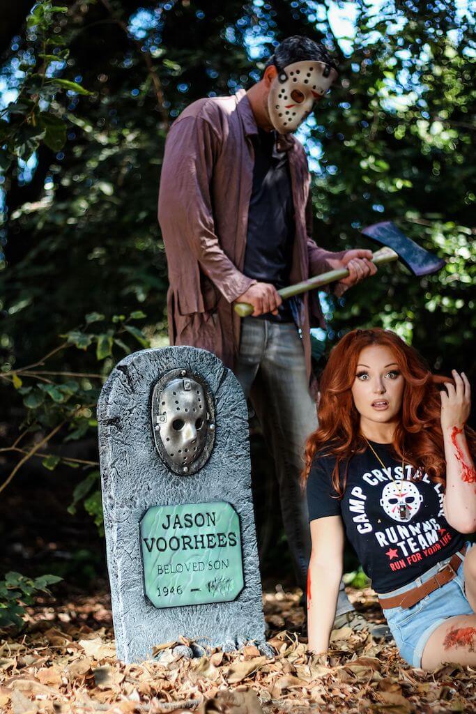 There is a girl with red hair, a black camp crystal lake running team shirt and denim shorts sitting in front of a Jason Voorhees tombstone, while he stands behind her in a hockey mask and brown shirt, while holding an axe.