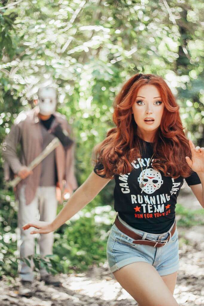There is a camp counselor with red hair, a black camp crystal lake running team shirt and denim shorts running, while Jason Voorhees stands behind her in a hockey mask and brown shirt, while holding an axe.