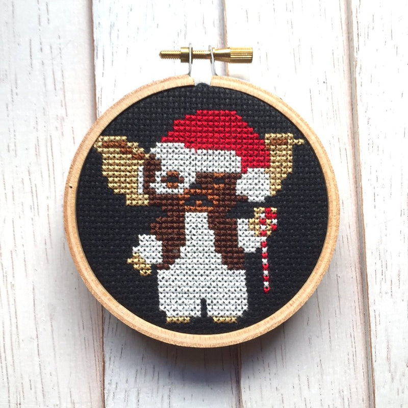 This is a Gremlins Gizmo cross stitch kit and it is black with a wooden circle hoop and he has on a red Santa hat and is holding a candy cane.