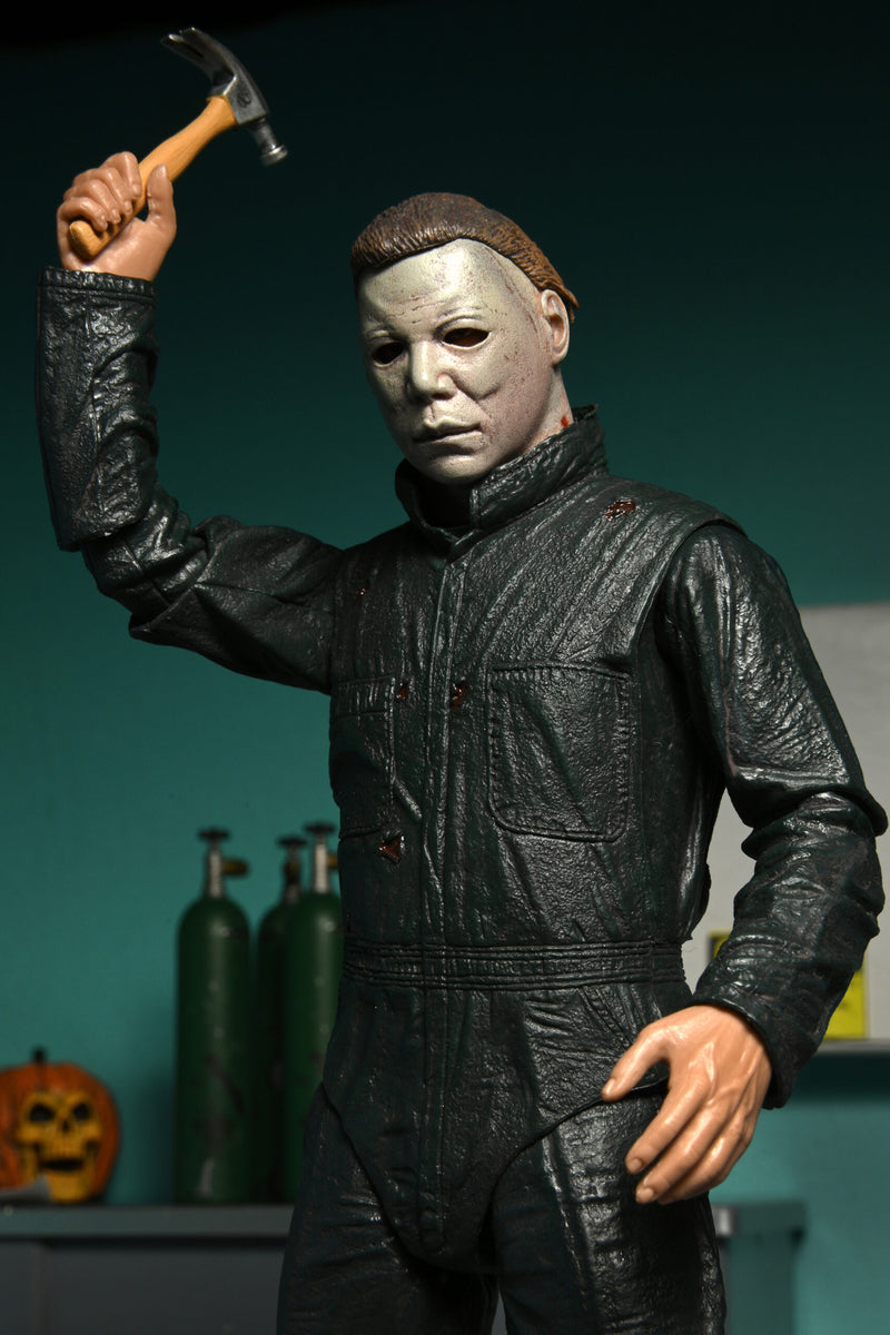 This is a NECA Halloween 2 Michael Myers and Loomis action figure set and Michael has a white mask, brown hair, green coveralls and is holding a silver hammer with brown handle, and there is an orange pumpkin.