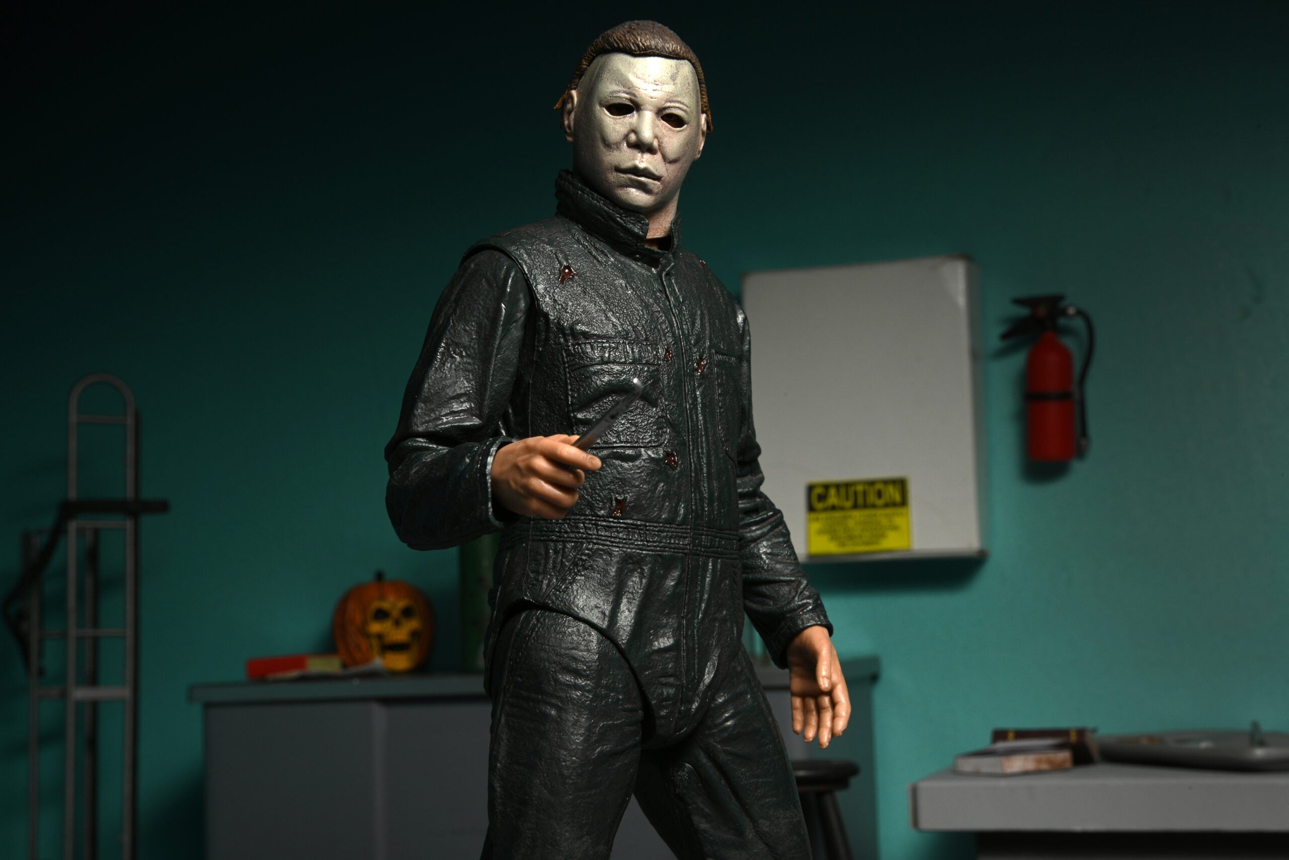 This is a NECA Halloween 2 Michael Myers and Loomis action figure set and Michael has a white mask, brown hair, green coveralls and is holding a silver scalpel and there is an orange pumpkin.