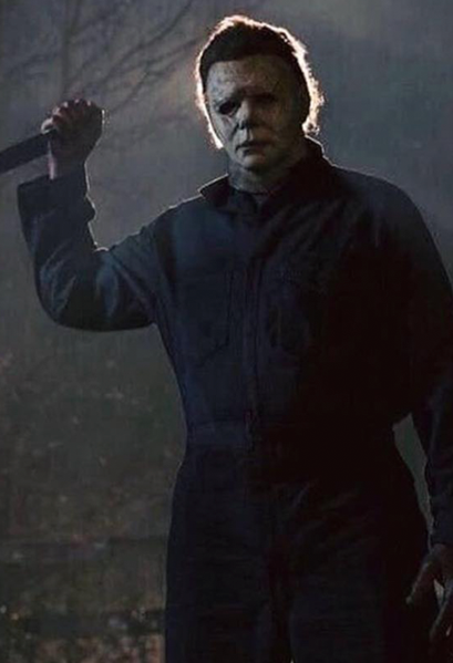 This is a Halloween 2018 Michael Myers mask that is a weathered grey face, neck and ear and has brown hair and black eyes and he is wearing grey coveralls, holding a knife and standing in front of a tree.