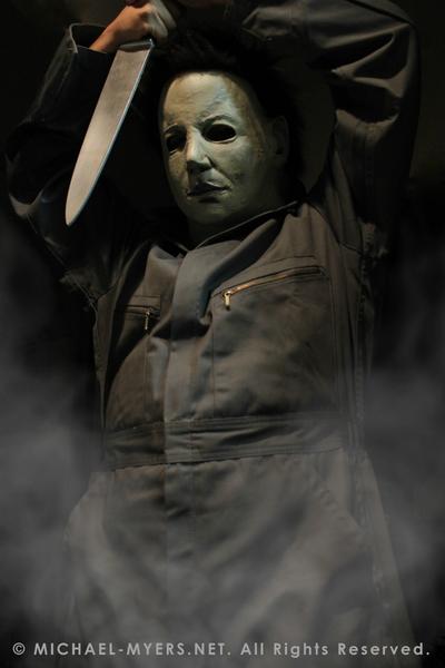 This is a Halloween 6 Curse of Michael Myers mask that is white with brown hair and black eyes and he is wearing coveralls and holding a knife above his head.