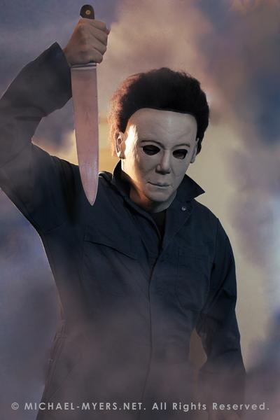 This is a Michael Myers Halloween H20 Mask that is white with brown hair and he is wearing green coveralls and holding a knife, while standing in smoke..