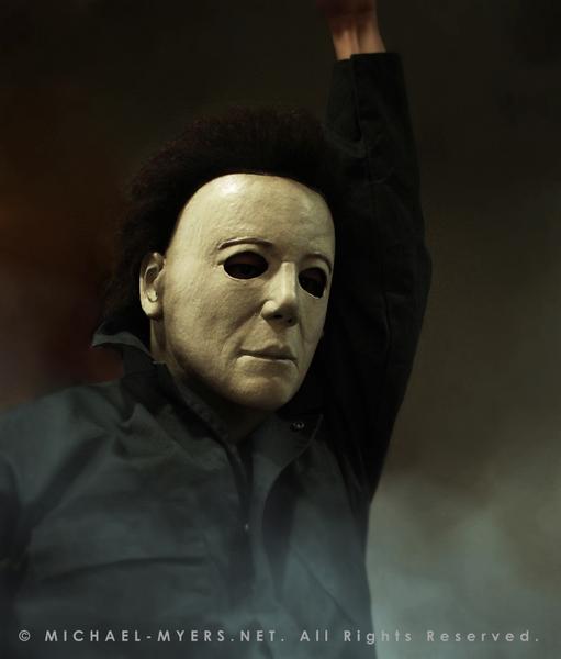 This is a Michael Myers Halloween H20 Mask that is white with brown hair and he is wearing green coveralls, while standing in smoke..