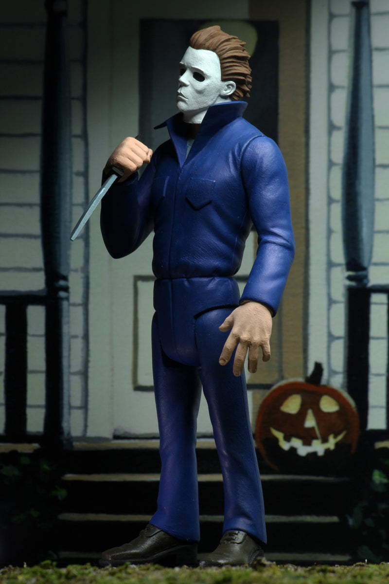 Michael Myers action figure is standing in blue coveralls on a front porch of a house in Haddonfield, holding a knife, with a pumpkin at his feet.