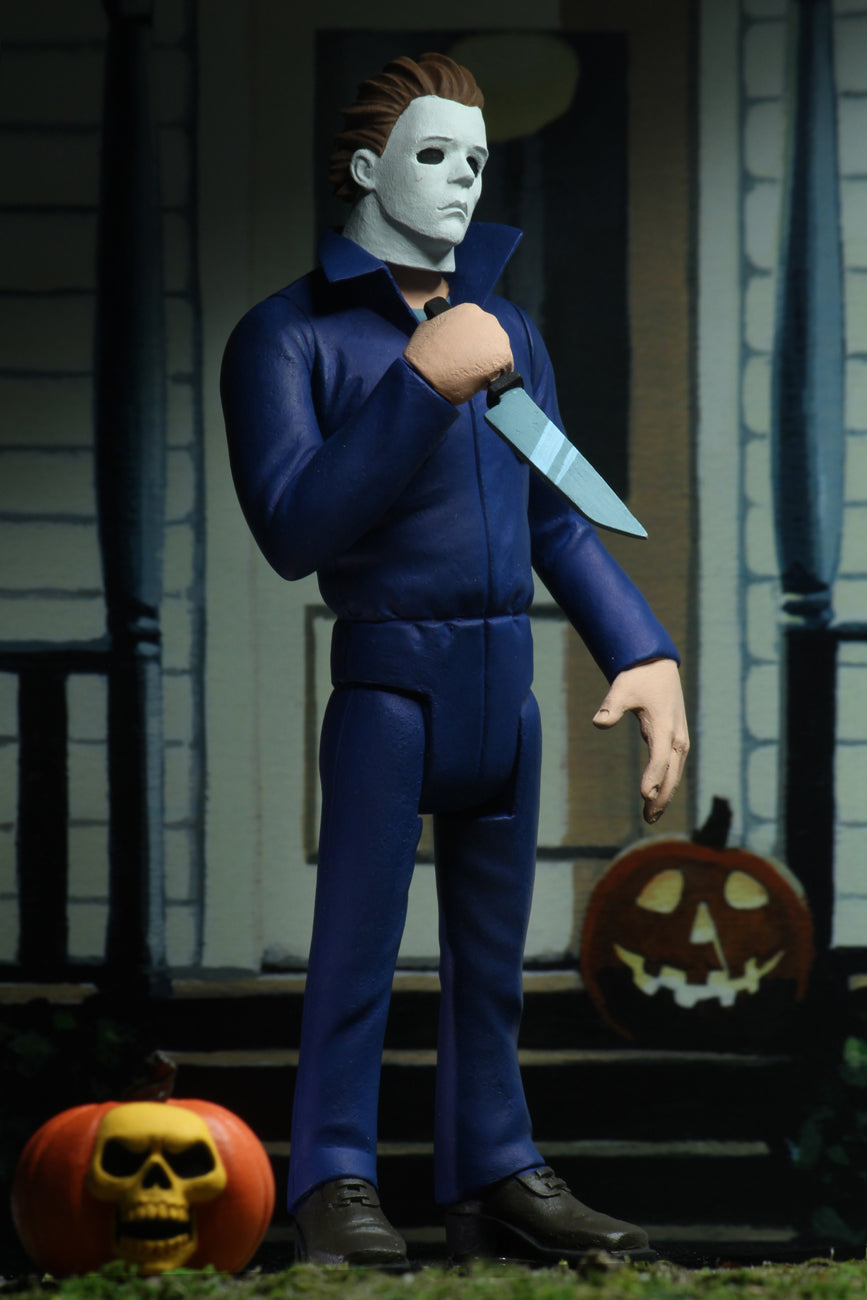 Michael Myers action figure is standing in blue coveralls on a front porch of a house in Haddonfield, holding a knife, with a pumpkin at his feet.