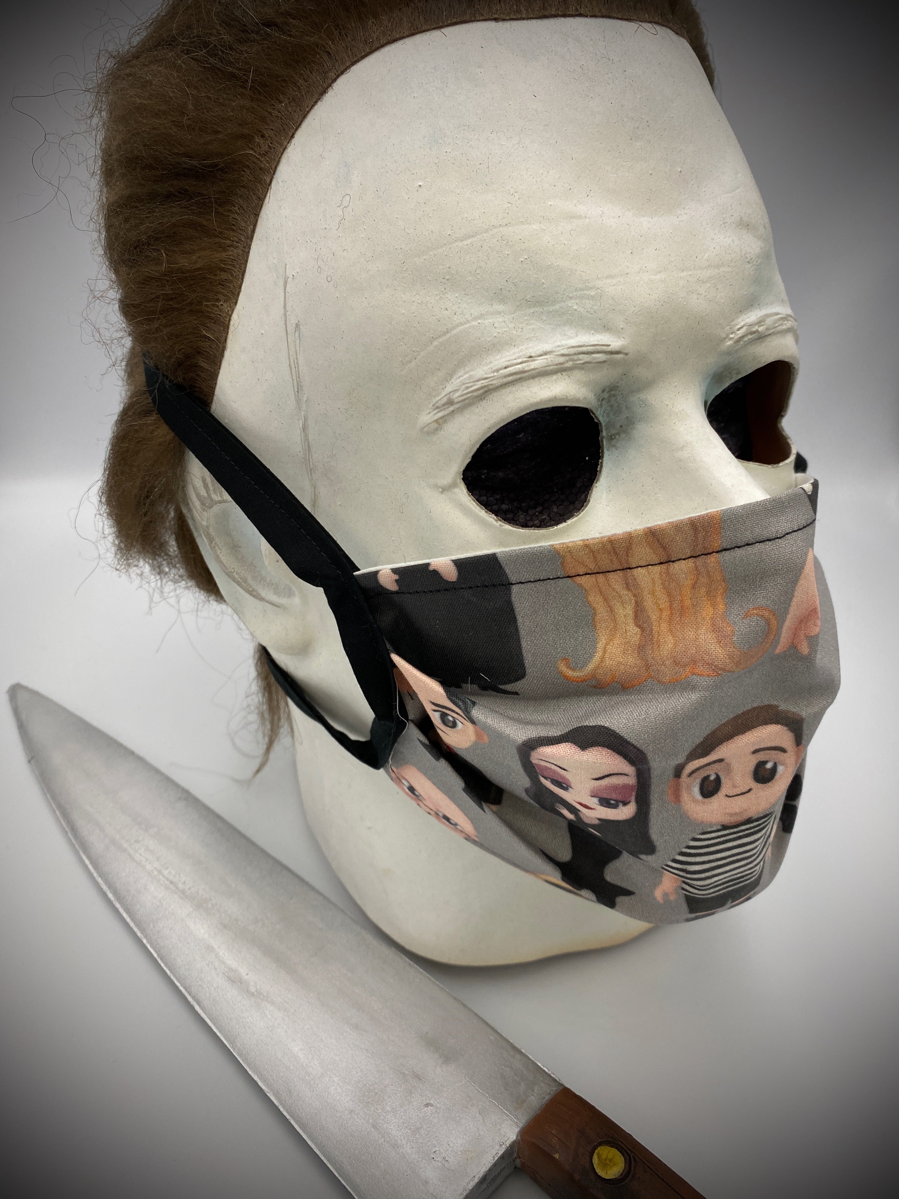This is a grey protective face mask with black ties and has Addams Family characters Gomez, Morticia, Wednesday, Pugsley, Lurch, Uncle Fester and Cousin Itt and is on a Michael Myers mask.