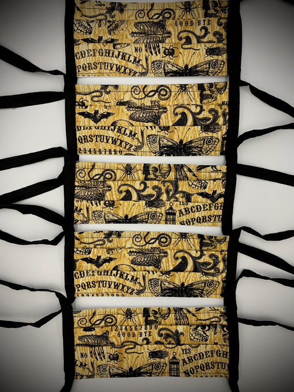 This is a brown protective face mask with black ties and it has a ouija board, skeleton, moth, bugs and cages.
