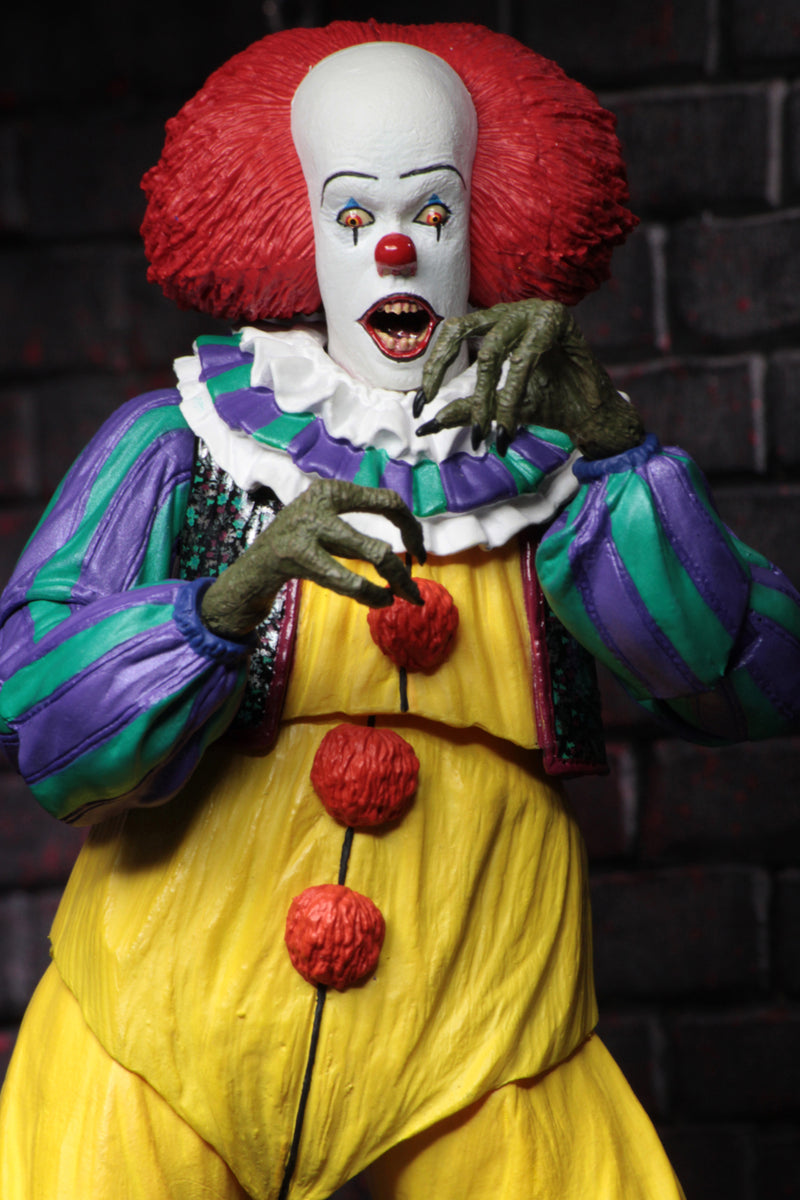 This is an It Movie 1990 NECA ultimate action figure with Pennywise who has a white face, red nose, orange red hair, yellow suit with orange balls and green and purple stripes and he has green claw hands.