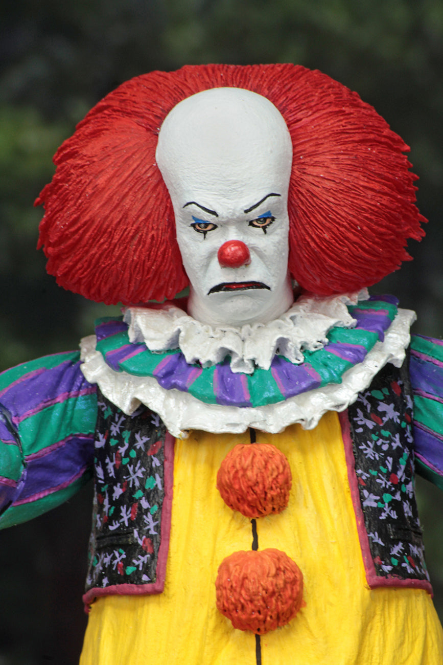 This is an It Movie 1990 NECA ultimate action figure with Pennywise who has a white face, red nose, orange red hair, yellow suit with orange balls and green and purple stripes with a flowered vest.