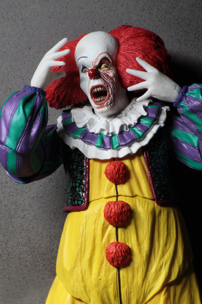 This is an It Movie 1990 NECA ultimate action figure with Pennywise who has a white face, red nose, orange red hair, yellow suit with orange balls and green and purple stripes and he is screaming and has a bloody face.
