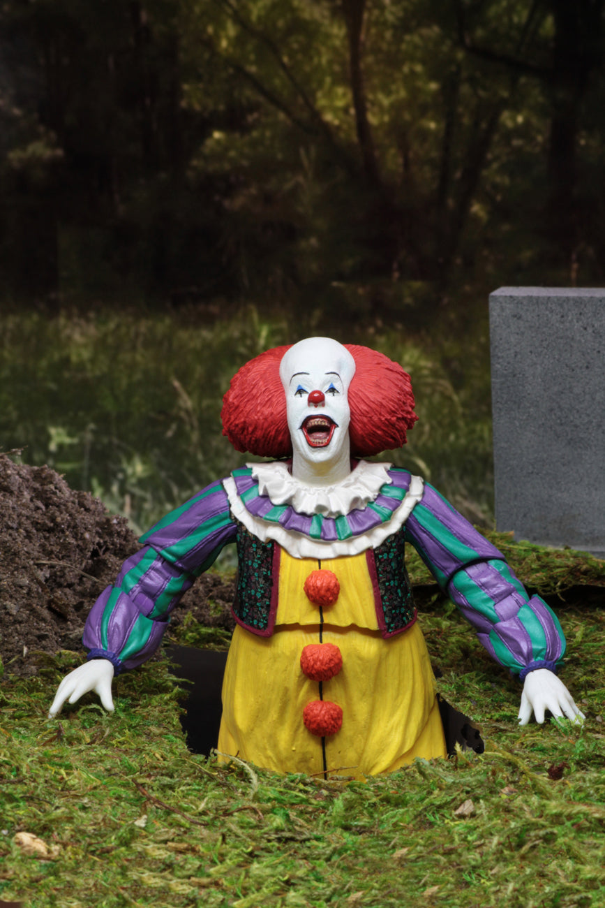 This is an It Movie 1990 NECA ultimate action figure with Pennywise who has a white face, red nose, orange red hair, yellow suit with orange balls and green and purple stripes and he is in a graveyard