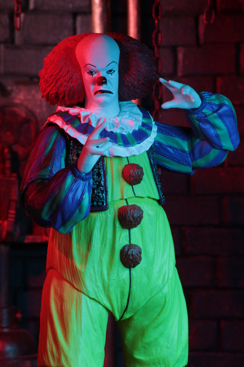 This is an It Movie 1990 NECA ultimate action figure with Pennywise who has a white face, red nose, orange red hair, yellow suit with orange balls and green and purple stripes, white gloves and he is in red light