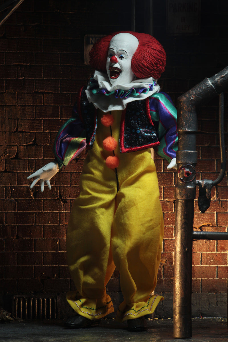 This is an It movie 1990 8' clothed action figure from NECA and Pennywise has a yellow suit with orange balls, purple and green striped sleeves, white face, yellow eyes, orange red hair, white gloves and he is smiling.