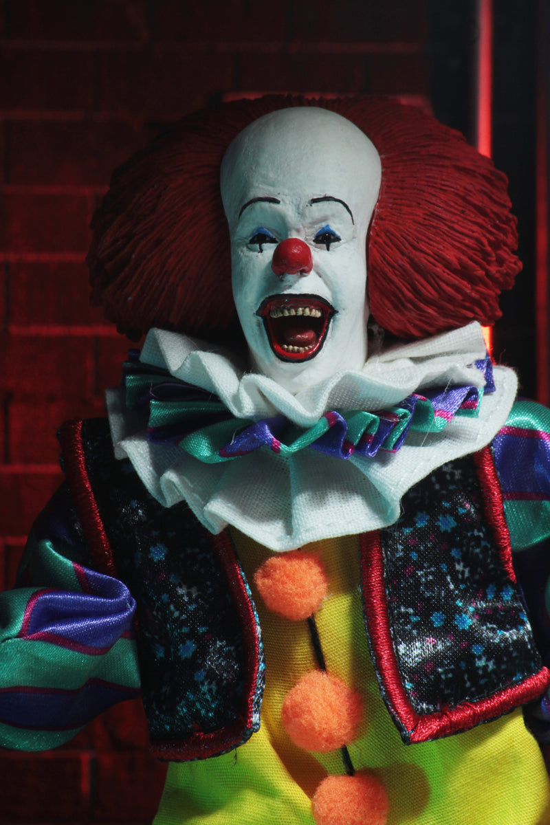 This is an It movie 1990 8' clothed action figure from NECA and Pennywise has a yellow suit with orange balls, purple and green striped sleeves, white face, yellow eyes, orange red hair, flowers on his vest and he is smiling