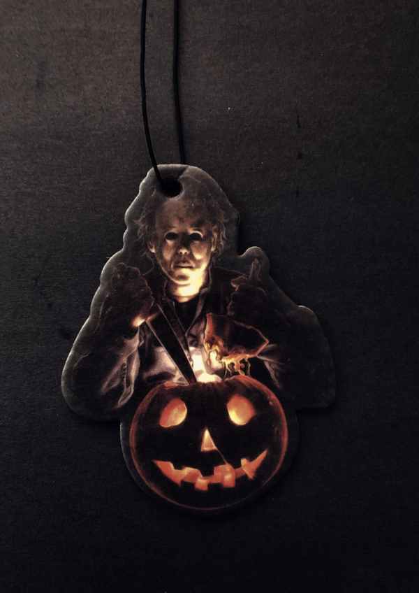 This is a Halloween movie Michael Myers air freshener and he has a white face, is holding a knife and an orange pumpkin that has a smile face.