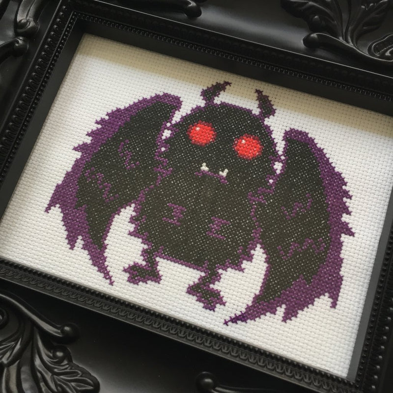 This is a Mothman DIY cross stitch kit and he is black and purple, with red eyes and white fangs.