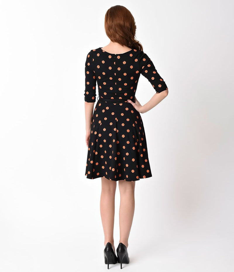 This is a black pinup flare dress with orange pumpkins, belt and 3/4 sleeves and the model is wearing black shoes and has brown hair to the side.
