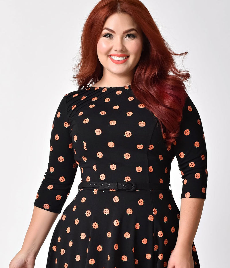This is a black pinup flare dress with orange pumpkins and 3/4 sleeves, belt and the plus model is smiling and has red hair.