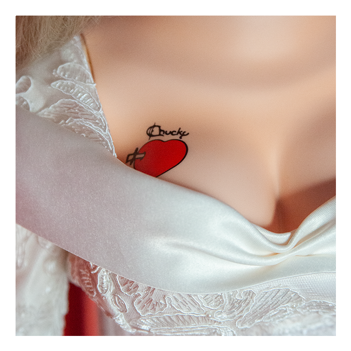 This is a Seed of Chucky Tiffany life size doll and she is wearing a white wedding dress and she has a red heart tattoo, with a knife through it.
