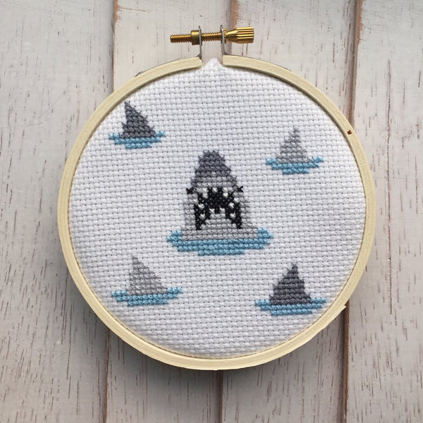 This is a DIY shark patterned cross stitch kit and they are grey fins, with blue water and he has pointy white teeth.