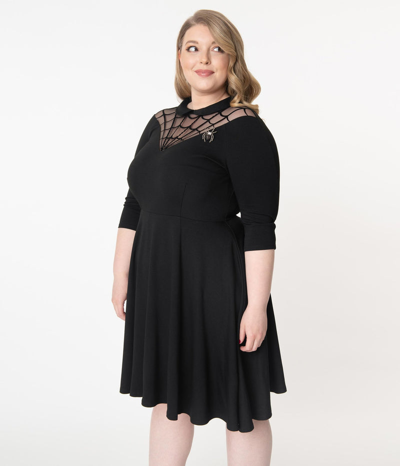 This is a black Unique Vintage flare dress that has a spiderweb neck, Peter Pan collar, 3/4 sleeves and the plus model is smiling and has a spider brooch. 