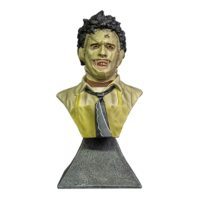 This is a Texas Chainsaw Massacre Leatherface mini bust and  he has brown hair, mask, yellow apron, tie and is on a grey stand.