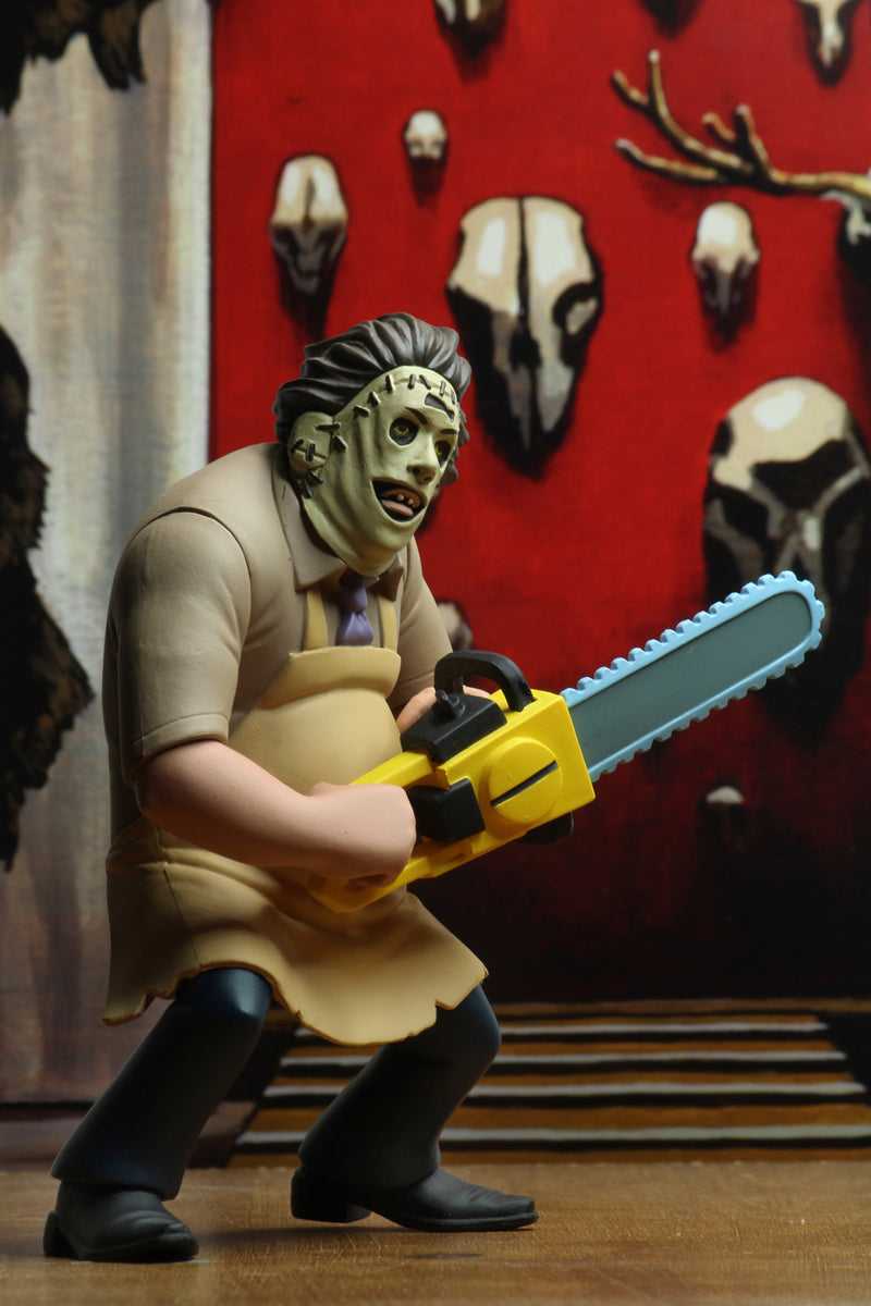Leatherface is standing in front of a red wall with animal skulls, while wearing a yellow apron and holding a yellow chainsaw.