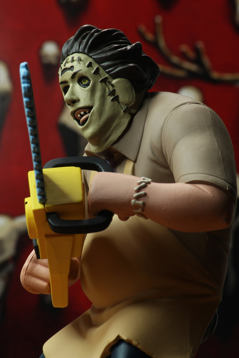Leatherface is standing in front of a red wall, while wearing a yellow apron and holding a yellow chainsaw.